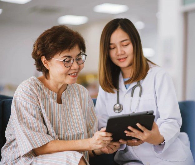 two people looking at a clipboard together. it appears to be a clinical setting, as the older person is wearing a hospital gown, and the younger person is wearing a white lab coat and stethoscope.