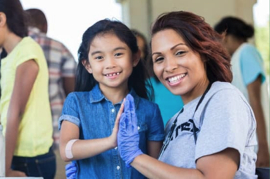 a photo of two people smiling. one is a young child with a band-aid, giving a high five to the other one, an adult wearing rubber gloves and a stethoscope