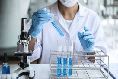 a photo of person wearing a lab coat, gloves, and a mask, and holding a test tube with blue liquid. there is a microscope to their right, and more test tubes are arranged in a rack.